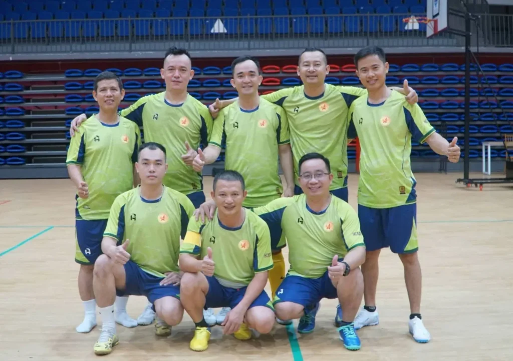Các thành viên đội bóng mặc áo xanh chụp ảnh tập thể tại sân bóng trong nhà.